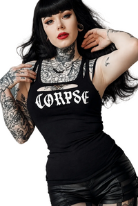 A tattooed woman with long black hair and red lipstick wears a black tank top with the word 'CORPSE' printed in white gothic lettering across the chest, posed against a plain light beige background.